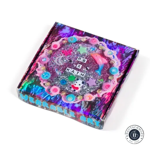Holografisch schimmernde Box „Wearable Art" der RB x KATM-Kollaboration mit glitzerndem Kranz-Motiv und Perlen, schräg fotografiert.