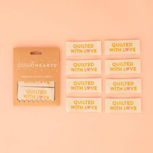 Quilted with Love – Verpackung Verpackung der Sarah Hearts Endfalt-Weblabels „Quilted with Love" in Metallic-Gold mit korallenrosa Herz