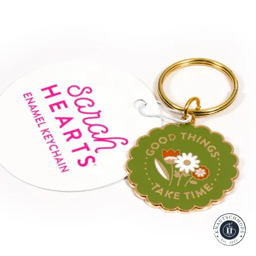 Sarah Hearts Emaille-Schlüsselanhänger „Good Things Take Time“ mit Blumenmotiv, neben der Anhängekarte.