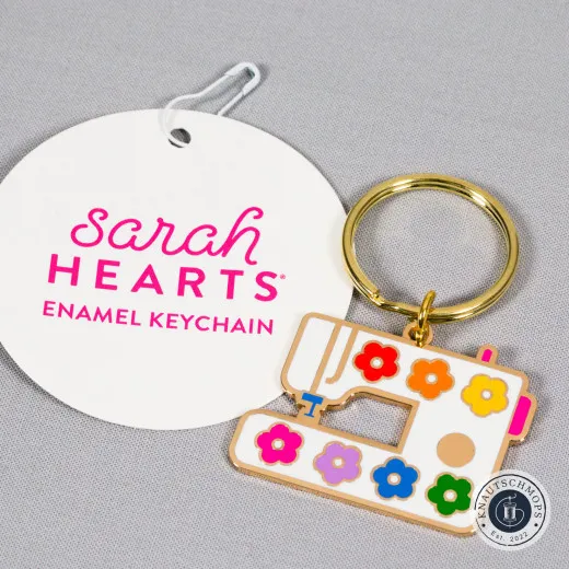 Sarah Hearts Emaille-Schlüsselanhänger in Nähmaschinenform mit bunten Blüten, neben der Anhängekarte.
