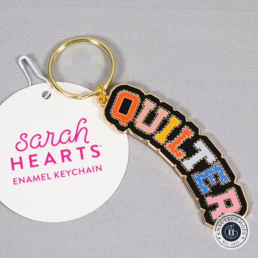 Sarah Hearts Emaille-Schlüsselanhänger „Quilter“ im College-Schriftzug, neben der Anhängekarte.
