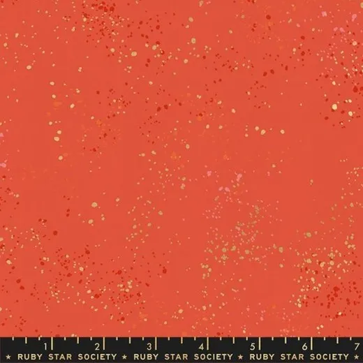 Nahaufnahme des melierten Baumwollstoffs Speckled in Festive mit goldmetallic- und rosafarbenen Farbspritzern auf korallrotem Grund, mit Maßband am Bildrand.