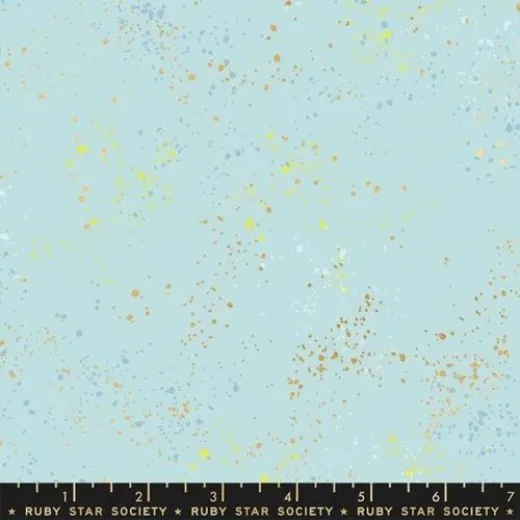 Nahaufnahme des melierten Baumwollstoffs Speckled in Polar mit goldmetallic-, gelben und hellblauen Farbspritzern auf zartem Eisblau, mit Maßband am Bildrand.