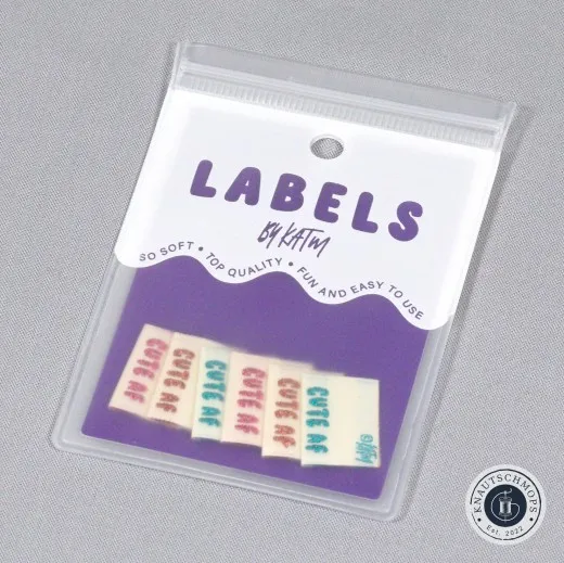 Verpackte Kylie and The Machine Labels „Cute AF" in glitzernden Metallic-Farben auf violetter Verpackungskarte.