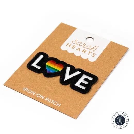 Verpackungskarte des Sarah Hearts Aufnähers „Love Pride" mit schwarzem Schriftzug „LOVE" und Regenbogen-Herz, in der Hand gehalten.