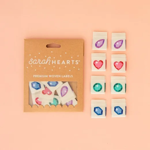 Sarah Hearts Verpackungskarton mit 8 Edelstein-Weblabels: lila Tropfen, pinkes Herz, türkiser Rundschliff und blauer Sechseck-Edelstein.