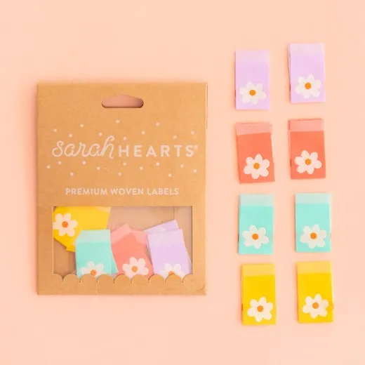 Verpackung mit 8 Daisy-Tag-Labels Kraftpapier-Verpackung „Daisy Tag" neben acht Weblabels mit weißem Gänseblümchen auf Lavendel, Koralle, Mint und Gelb.