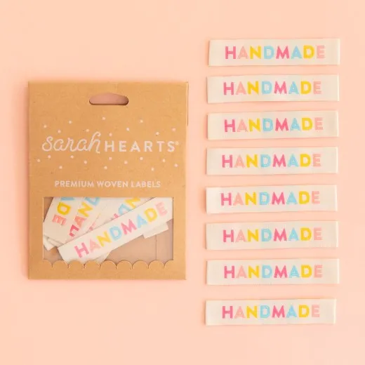 Kraftpapier-Verpackung „Handmade" neben acht cremefarbenen Weblabels mit dem Schriftzug „HANDMADE" in Regenbogenfarben.