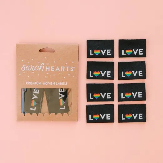 Verpackung mit 8 Love-Pride-Labels Kraftpapier-Verpackung „Love Pride" neben acht schwarzen Weblabels mit weißem „LOVE"-Schriftzug und Regenbogenherz.