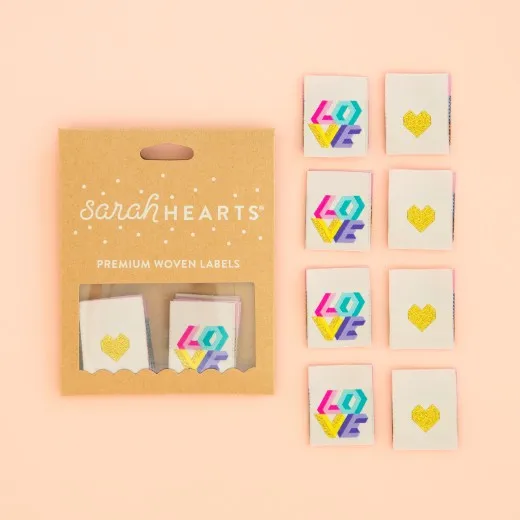 Verpackung mit 8 Love-Quilt-Labels Kraftpapier-Verpackung „Love Quilt" neben acht cremefarbenen Weblabels mit geometrischem „LOVE"-Schriftzug in Pink, Türkis, Lila und Gelb.