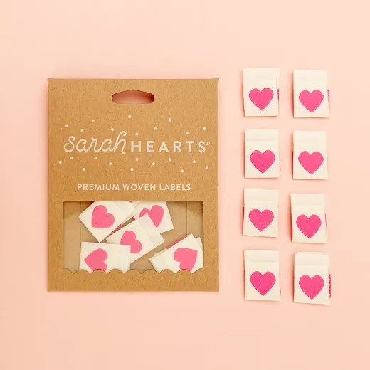 Verpackung mit 8 Pink-Heart-Labels Kraftpapier-Verpackung „Pink Heart" neben acht cremefarbenen Weblabels mit pinkfarbenem Herz.