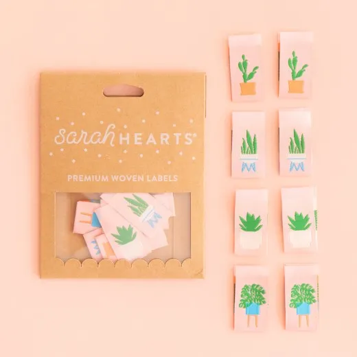 Verpackung mit 8 Zimmerpflanzen-Labels Kraftpapier-Verpackung „Houseplants" neben acht rosafarbenen Weblabels mit Kaktus, Grasähre, Sukkulente und Monstera im Topf.