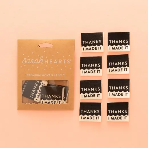 Verpackung mit vollständigem 8er-Set Sarah-Hearts-Weblabel „Thanks I Made It" in Kraftpapier-Verpackung neben allen acht schwarzen Labels mit weißem Siebdruck.