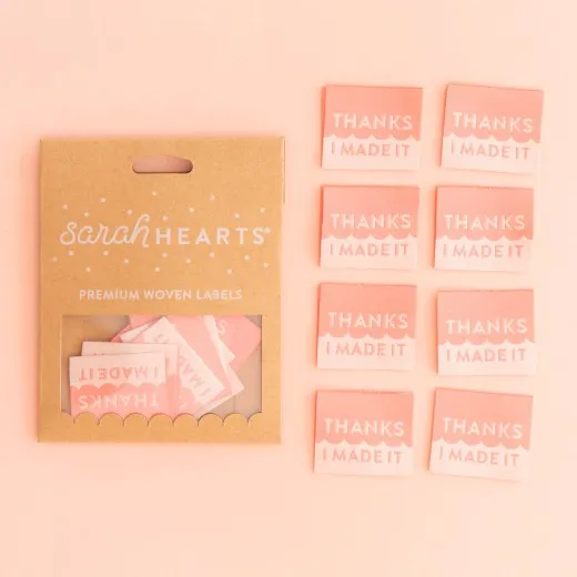 Verpackung mit vollständigem 8er-Set Sarah-Hearts-Weblabel „Thanks I Made It" in Kraftpapier-Verpackung neben allen acht Labels in Koralle und Creme.