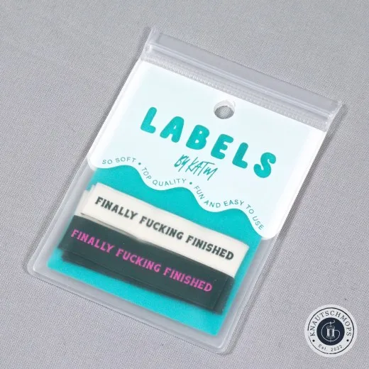 „Finally Fucking Finished"-Labels in Creme/Dunkelgrün und Waldgrün/Pink, sichtbar in der Original-Verpackungskarte „Labels by KATM", schräg fotografiert.