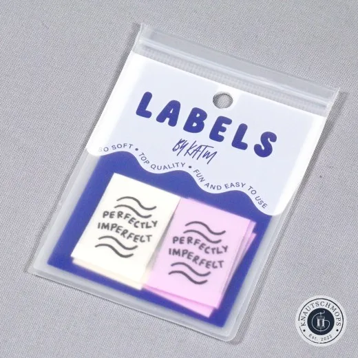 „Perfectly Imperfect"-Labels in Elfenbein und Flieder, sichtbar in der Original-Verpackungskarte „Labels by KATM", schräg fotografiert.