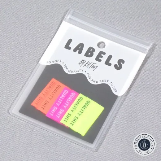 Verpackung schräg fotografiert Sechs „Quality Shit"-Labels in Neongelb, Neonorange und Knallpink, sichtbar in der Original-Verpackungskarte „Labels by KATM", schräg fotografiert.