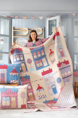 Frau präsentiert einen fertig genähten Patchwork-Quilt mit Dorfmotiv aus der Kollektion Village Life, dazu passende Kissen im Vordergrund.