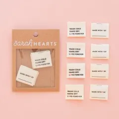 Vier „Wash Cold, Hang Dry, Love Forever“- und vier „Made With Love By“-Labels ausgelegt auf rosa Hintergrund.