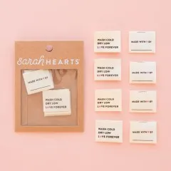 Vier „Wash Cold, Dry Low, Love Forever“- und vier „Made With Love By“-Labels ausgelegt auf rosa Hintergrund.