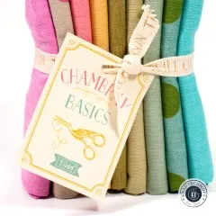 Nahaufnahme des Papieranhängers „Chambray Basics" mit türkisfarbenem Tilda-Logo neben Pink- und Taupe-Stoffkanten.