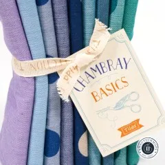 Nahaufnahme des Papieranhängers „Chambray Basics" mit blauem Tilda-Logo neben Lila- und Blau-Stoffkanten.