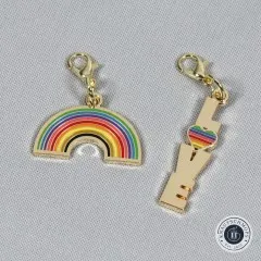 Regenbogen- und 'Love'-Anhänger nebeneinander auf grauem Stoff.