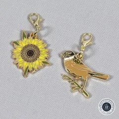 Vogel- und Sonnenblumen-Anhänger nebeneinander auf grauem Stoff.