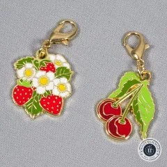 Erdbeer- und Kirsch-Anhänger nebeneinander auf grauem Stoff, schräge Perspektive.