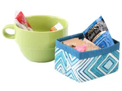 Kleiner blauer Stoffkorb mit Rauten-Muster, gefüllt mit Teebeuteln, neben grüner Kaffeetasse.
