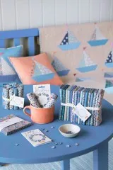 Deko-Ansicht: Segelboot-Kissen und -Quilt sowie gerollte Stoffe der Summer-Kollektion auf blauem Tisch.
