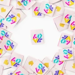 Ein cremefarbenes Weblabel mit geometrischem „LOVE"-Schriftzug mittig platziert, umgeben von locker verstreuten Labels – teils mit Vorderseite, teils mit goldenem Herz auf der Rückseite sichtbar.
