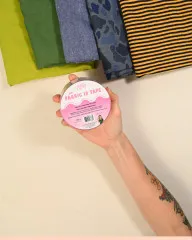 Hand hält die Fabric-ID-Tape-Rolle vor gefalteten Stoffen in Grün, Blau und Senfgelb