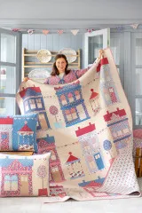 Frau präsentiert einen fertig genähten Patchwork-Quilt mit Dorfmotiv aus der Kollektion Village Life, dazu passende Kissen im Vordergrund.