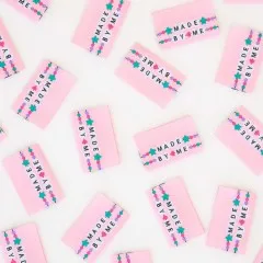 Weitere Nahaufnahme der rosa „Made by Me"-Weblabels mit Perlen-, Stern- und Herzmotiv.