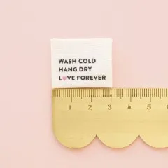 Wash-Pflegeetikett mit Herzsymbol neben einem goldenen Lineal zum Größenvergleich.