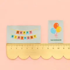 Happy-Birthday- und Ballon-Label neben einem goldenen Lineal zum Größenvergleich.