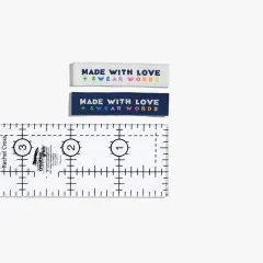 Je ein „Made With Love & Swear Words"-Label in Creme und Marineblau neben einem Lineal zum Größenvergleich.
