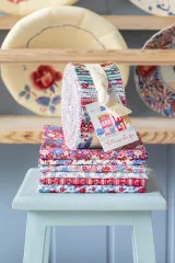 Tilda Fabric Jelly Roll auf gefalteten Stoffen stehend, dekorativ vor einem Regal mit Geschirr arrangiert.