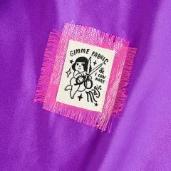 Das Label „Gimme fabric & I can make magic“ mit Scheren schwingender Figur ist auf lila Satinstoff aufgenäht, umrahmt von einem ausgefransten pink-silbernen Stoffflicken.