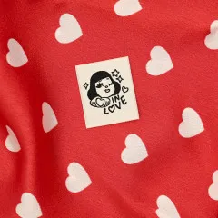 Das Label „In Love“ mit einer Herz haltenden Figur ist auf roten Stoff mit weißem Herzchen-Muster aufgenäht.