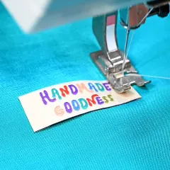 'Handmade Goodness'-Label wird an der Nähmaschine auf türkisfarbenen Stoff genäht