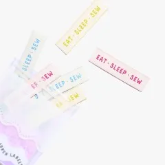 Labels 'Eat Sleep Sew' in Rosa, Blau und Gelb beim Herausfallen aus der Verpackung.