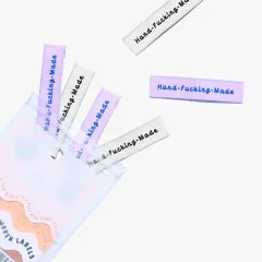 Labels 'Hand-Fucking-Made' in Flieder und Creme beim Herausfallen aus der Verpackung.