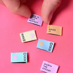 Mehrere Mini-Labels verstreut auf rosa Stoff, eines wird zwischen den Fingern gehalten.