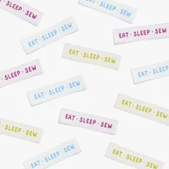 Mehrere Labels 'Eat Sleep Sew' in drei Farben verstreut in der Draufsicht.
