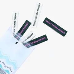 Mehrere „Finally Fucking Finished"-Labels in Creme und Waldgrün lose verstreut, teils aus der geöffneten Verpackung ragend.