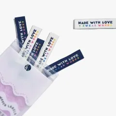 Mehrere „Made With Love & Swear Words"-Labels in Marineblau und Creme lose verstreut, teils aus der geöffneten Verpackung ragend.