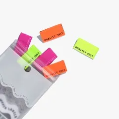 Mehrere „Quality Shit"-Labels in Neongelb, Neonorange und Knallpink lose verstreut, teils aus der geöffneten Verpackung ragend.