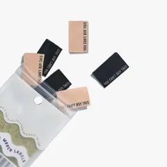 Mehrere „You Can't Buy This"-Labels in Schwarz und Kamel lose verstreut, teils aus der geöffneten Verpackung ragend.